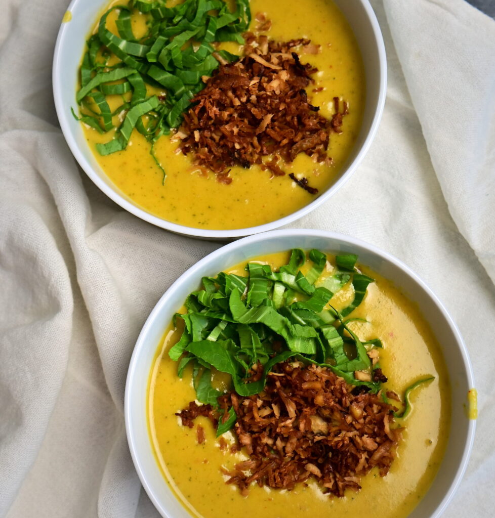 Two white bowls of creamy yellow potato soup with vegan bacon and greens, served on a white placemat.