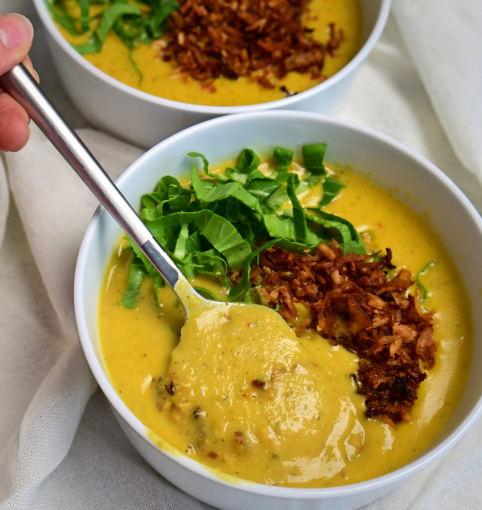 Two white bowls of creamy yellow potato soup with vegan bacon and greens on a white cloth, a spoon holding some soup.