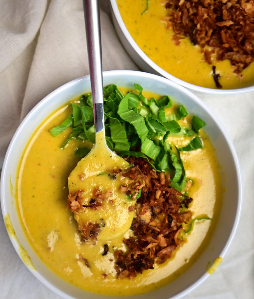 Two white bowls of creamy yellow potato soup with vegan bacon and greens on a white cloth, a spoon holding some soup.
