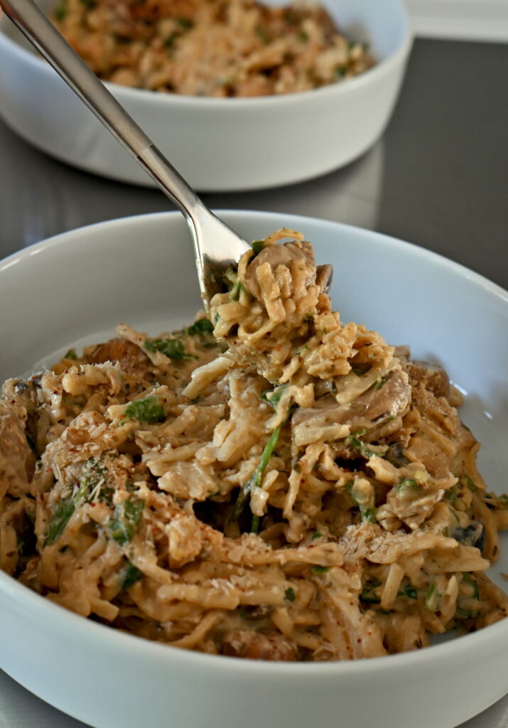 a white bowl and a fork lifting with gf rice noodles with mushroom sauce, herbs, creamy, with toppings - walnuts, nooch chili flakes