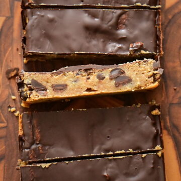 chickpea Protein Cookie Dough bars covered with chocolate (one's side facing the camera) photographed from top and placed on a wooden cutting board