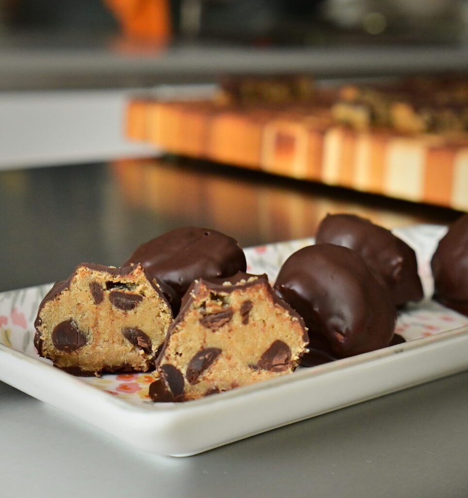 chickpea Protein Cookie Dough balls covered with chocolate on a flowered plate on a metal surface, one ball cut open