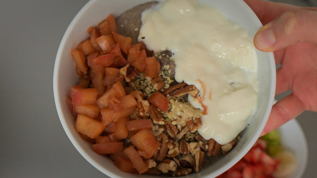hand holding white bowl of oats with vegan soy yoghurt, pecans, caramelized cinnamon apples and fruits