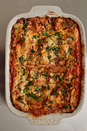 vegan lasagna with cashew bechamel and red zucchini + ground meat sauce in a red pan with basil leaves