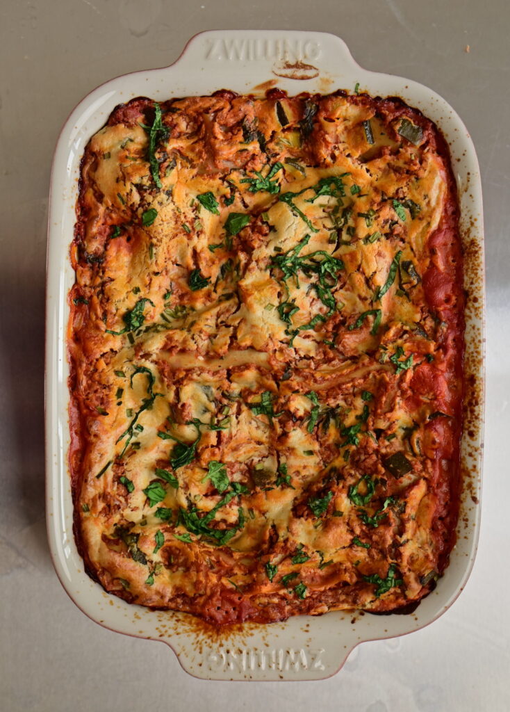 vegan lasagna with cashew bechamel and red zucchini + ground meat sauce in a red pan with basil leaves