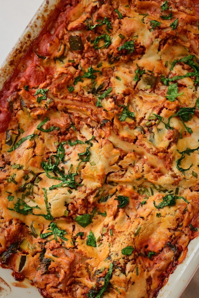 vegan lasagna with cashew bechamel and red zucchini + groung meat sauce in a red pan with basil leaves
