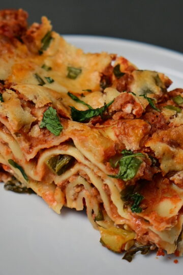 a slice of vegan lasagna with cashew bechamel and red zucchini + ground meat sauce with basil leaves, served on a white plate on a metal surface