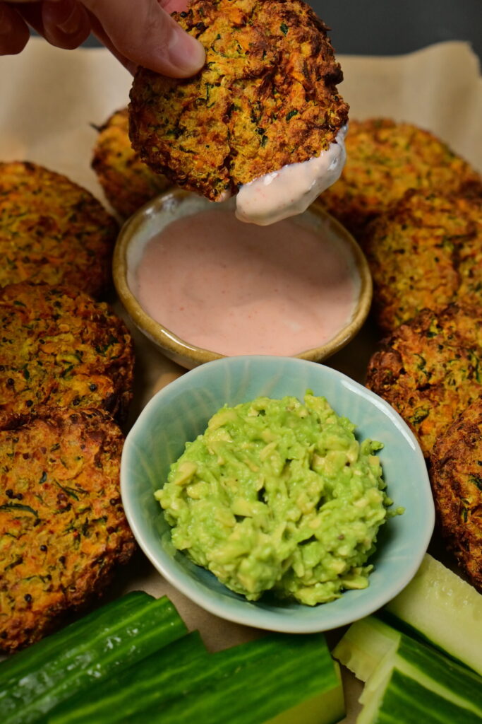 hand dipping a carrot and zucchini fritter in a orange deep next to guacamole, cucumbers and other fritters