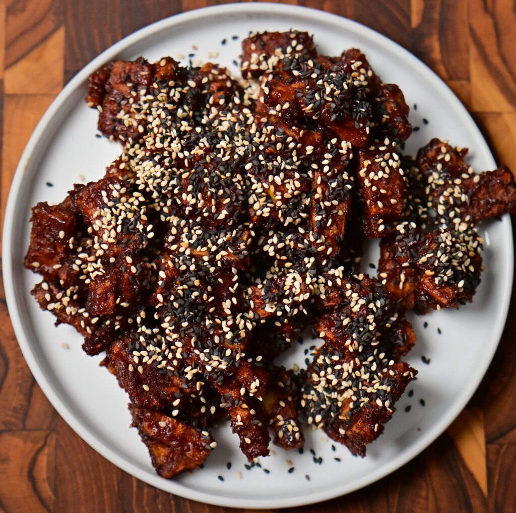 a white plate with sweet and spicy sticky tofu with sesame placed on a wooden board