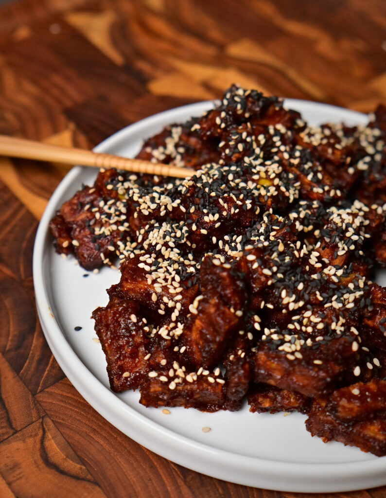 a white plate with sweet and spicy sticky tofu with sesame placed on a wooden board