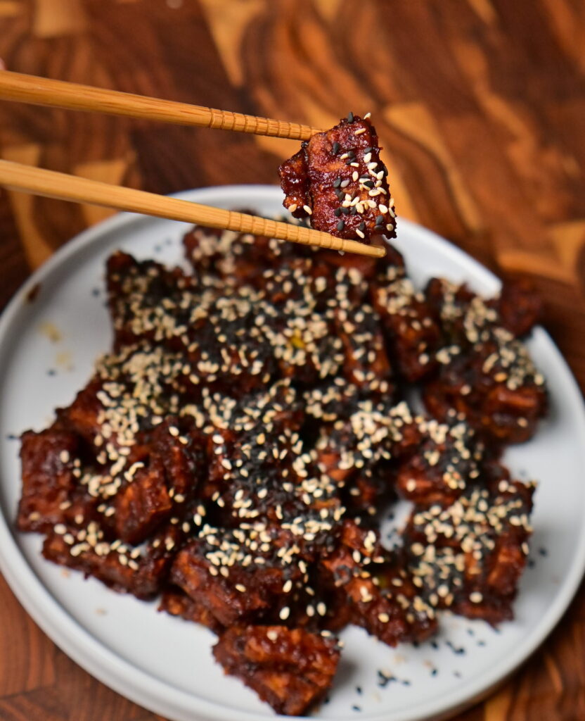 chop sticks hold sweet and spicy sticky tofu with sesame placed on a wooden board