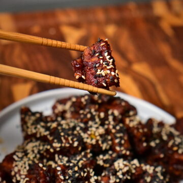 chop sticks hold sweet and spicy sticky tofu with sesame placed on a wooden board