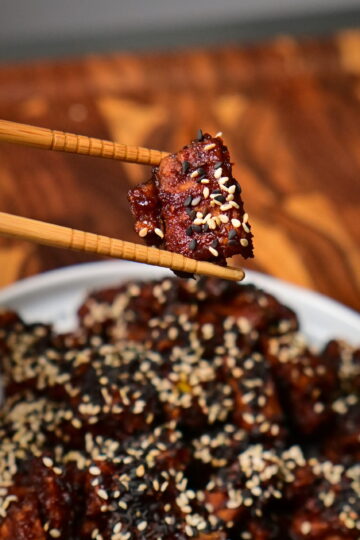 chop sticks hold sweet and spicy sticky tofu with sesame placed on a wooden board