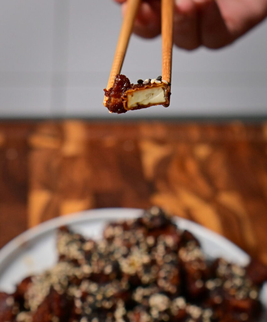 chop sticks hold sweet and spicy sticky tofu with sesame placed on a wooden board