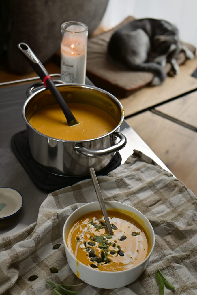 silky bright orange vegetables soup in a pot with a ladle, bowl. and a candle and dog in the background.