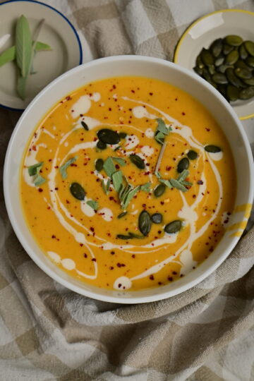 silky bright orange vegetables soup in a bowl, topped with pumpkin seeds, cream, sage and chilli.