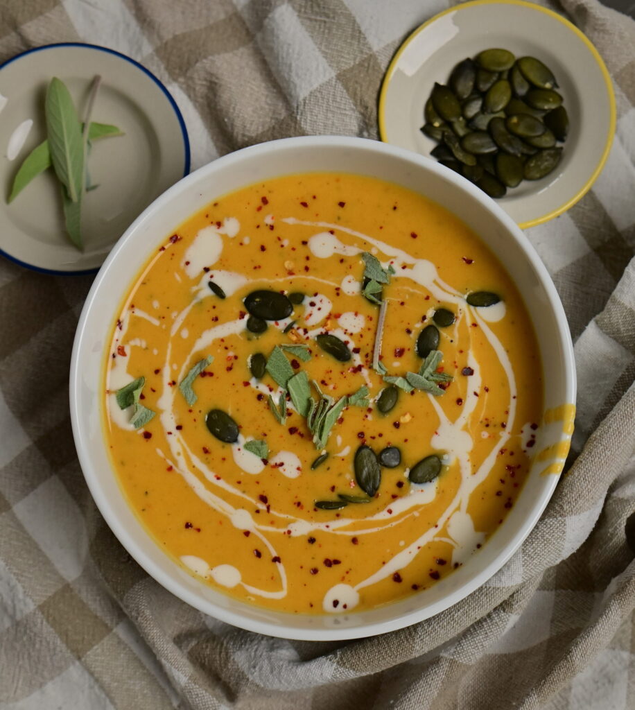 silky bright orange vegetables soup in a bowl, topped with pumpkin seeds, cream, sage and chilli.