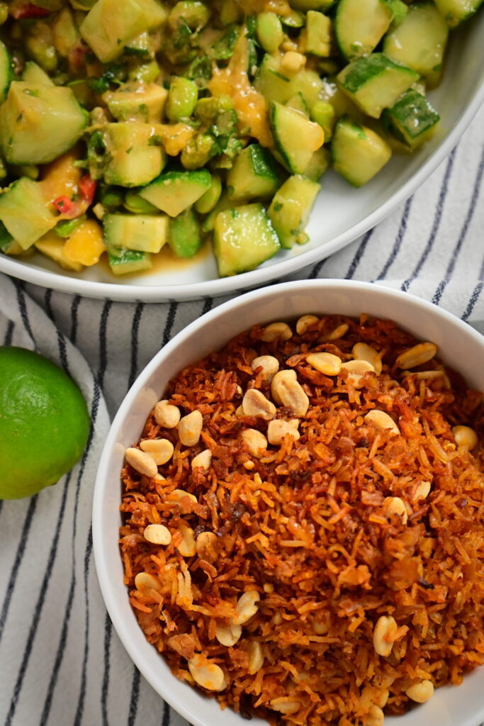 A white bowl with Thai-style salad and a wooden spoon, a white bowl with crispy rice, tofu, and peanuts, a striped tablecloth, peanut–lime dressing.