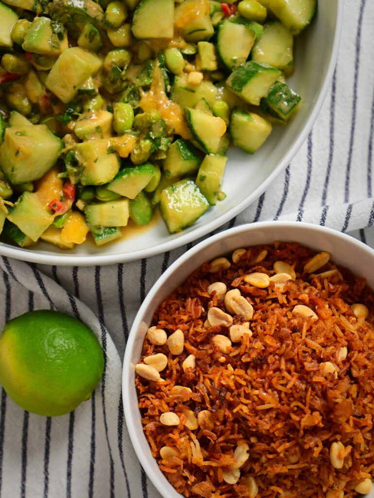 A white bowl with Thai-style salad and a wooden spoon, a white bowl with crispy rice, tofu, and peanuts, a striped tablecloth, peanut–lime dressing.