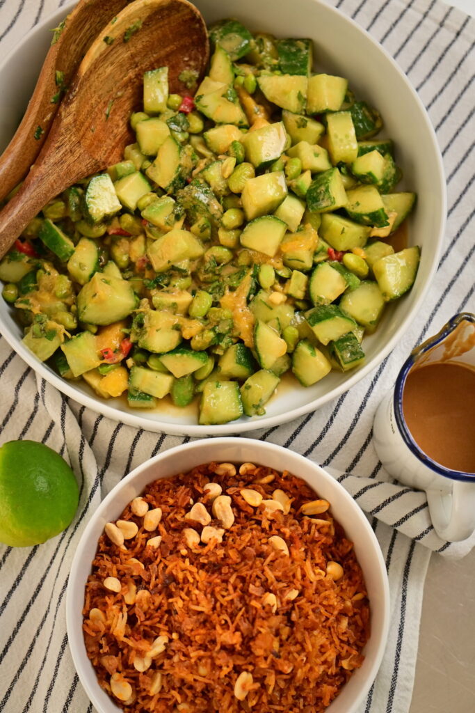 A white bowl with Thai-style salad and a wooden spoon, a white bowl with crispy rice, tofu, and peanuts, a striped tablecloth, peanut–lime dressing.