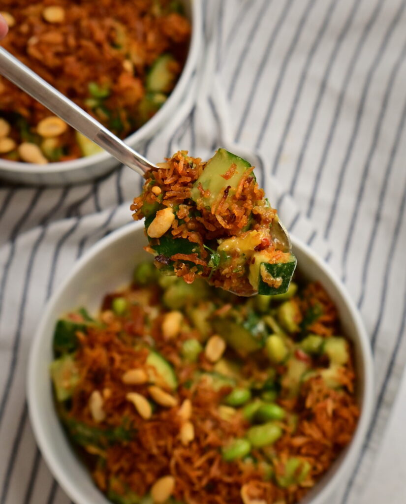 A spoon holding Thai-style crispy rice salad above two white bowls with the salad, on a white striped tablecloth.