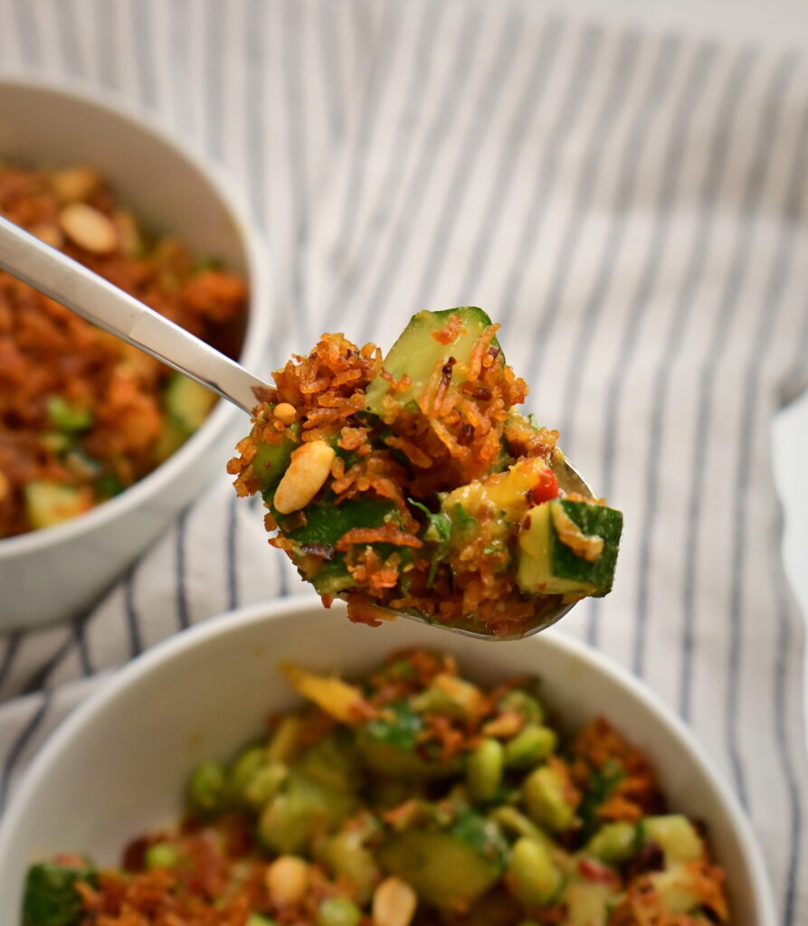 A spoon holding Thai-style crispy rice salad above two white bowls with the salad, on a white striped tablecloth.