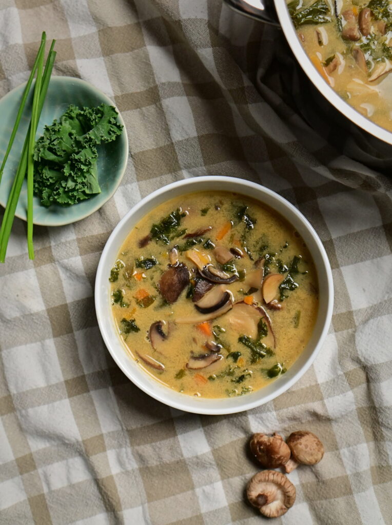 White bowl of mushroom soup with carrots and greens, beside herbs, shiitake mushrooms, and poppy seed challah on a brown and white kitchen towel