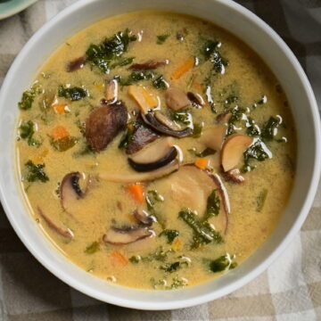 White bowl of mushroom soup with carrots and greens, beside herbs, shiitake mushrooms, and poppy seed challah on a brown and white kitchen towel