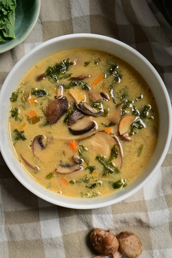 White bowl of mushroom soup with carrots and greens, beside herbs, shiitake mushrooms, and poppy seed challah on a brown and white kitchen towel