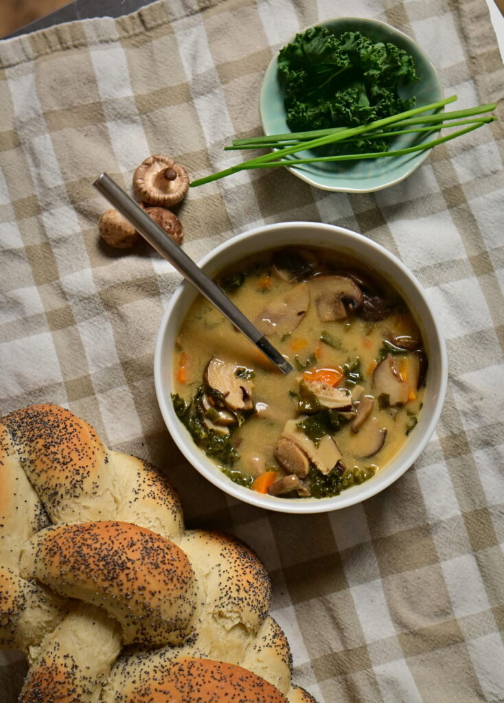 White bowl of mushroom soup with carrots and greens, beside herbs, shiitake mushrooms, and poppy seed challah on a brown and white kitchen towel