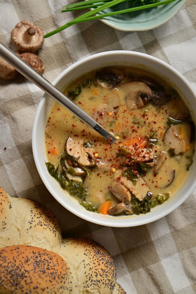 White bowl of mushroom soup with carrots and greens, beside herbs, shiitake mushrooms, and poppy seed challah on a brown and white kitchen towel