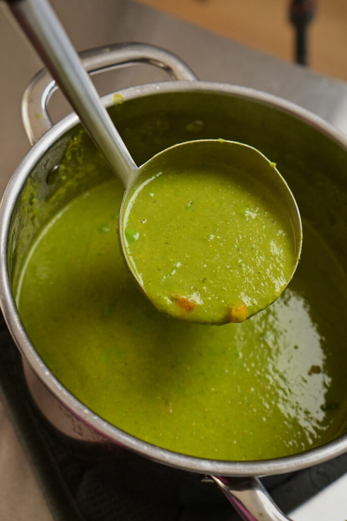 Stainless steel pot with thick green creamy pea soup with a ladle with soup and carrots