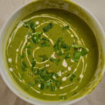 Bowl of creamy pea soup drizzled with cream and topped with herbs.