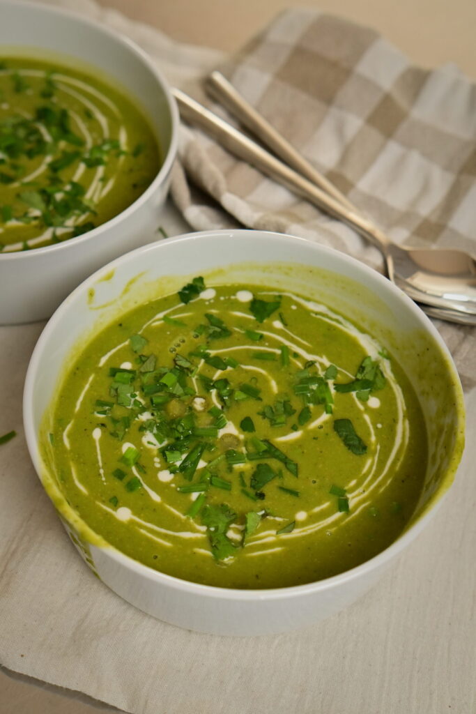 Two white bowls of creamy pea soup drizzled with cream and herbs, one bowl in the foreground.