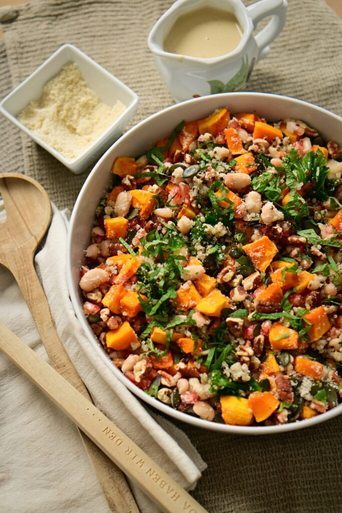 A serving bowl with kale salad, serving spoons, almond flour, on a brown tablecloth and a cream-colored cloth napkin