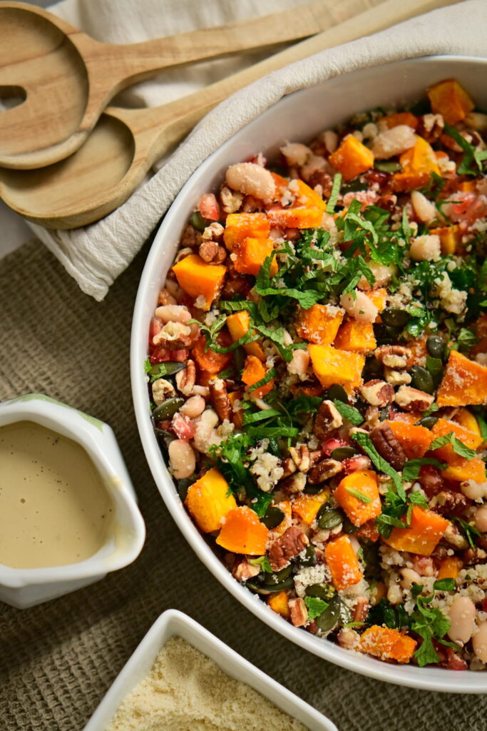 A serving bowl with kale salad, serving spoons, almond flour, on a brown tablecloth and a cream-colored cloth napkin