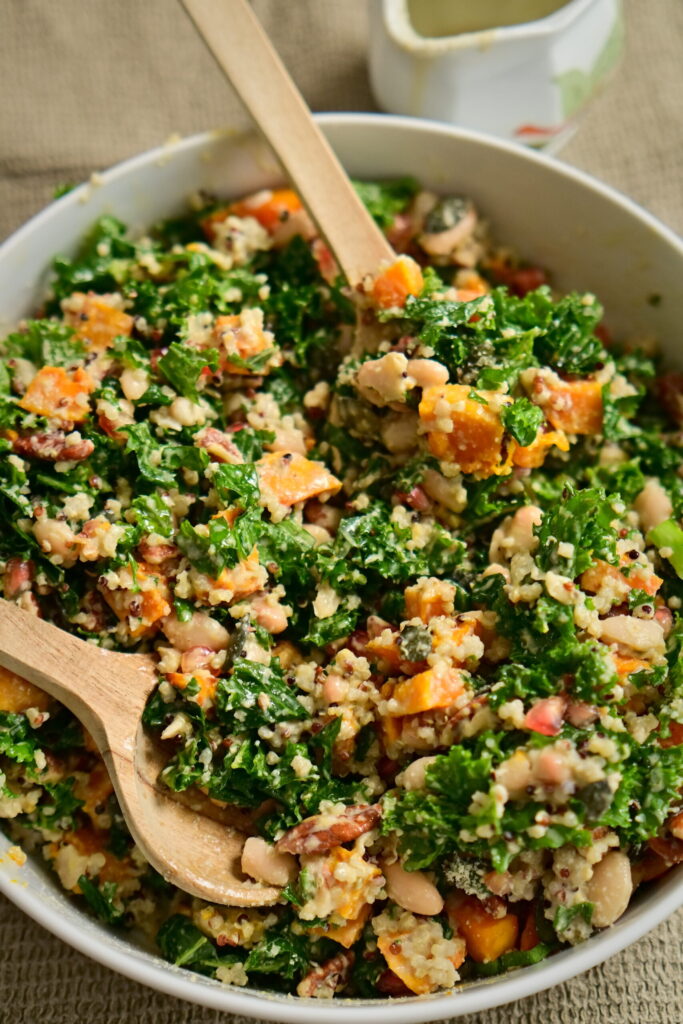 A serving bowl with kale salad, serving spoons, almond flour, on a brown tablecloth and a cream-colored cloth napkin