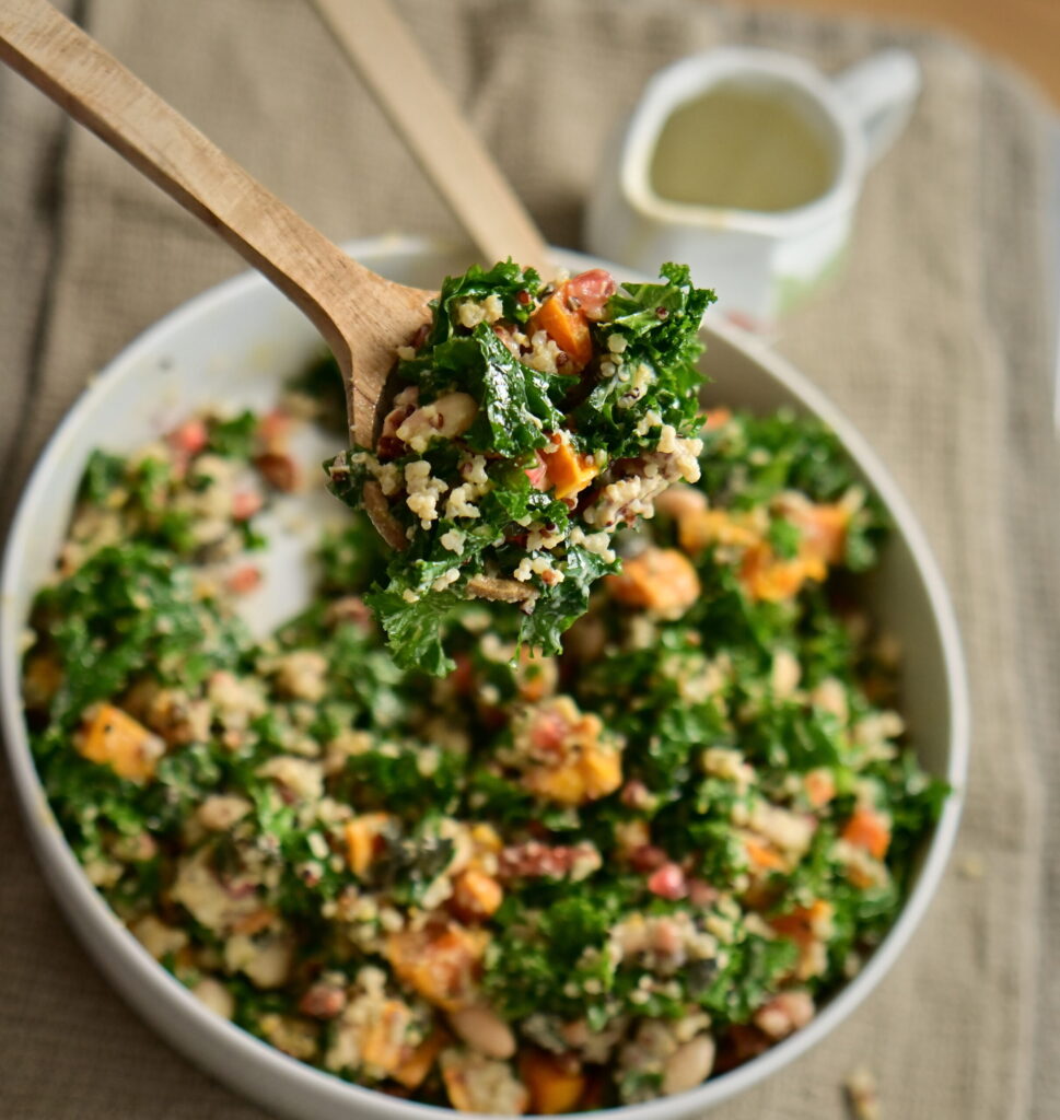 An overhead serving spoon holding kale and pumpkin salad, with a serving bowl below and an elegant pitcher for the dressing