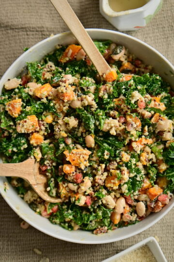 A serving bowl with kale and pumpkin salad, serving spoons, a pitcher, and almond flour on a brown tablecloth