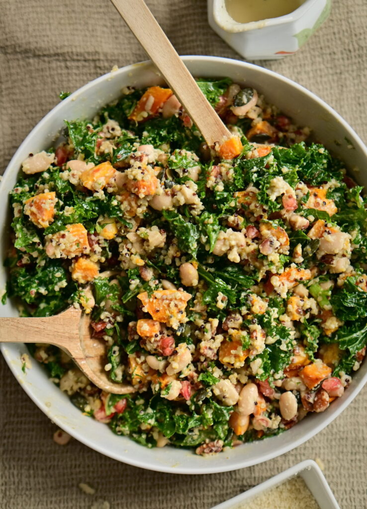 A serving bowl with kale and pumpkin salad, serving spoons, a pitcher, and almond flour on a brown tablecloth