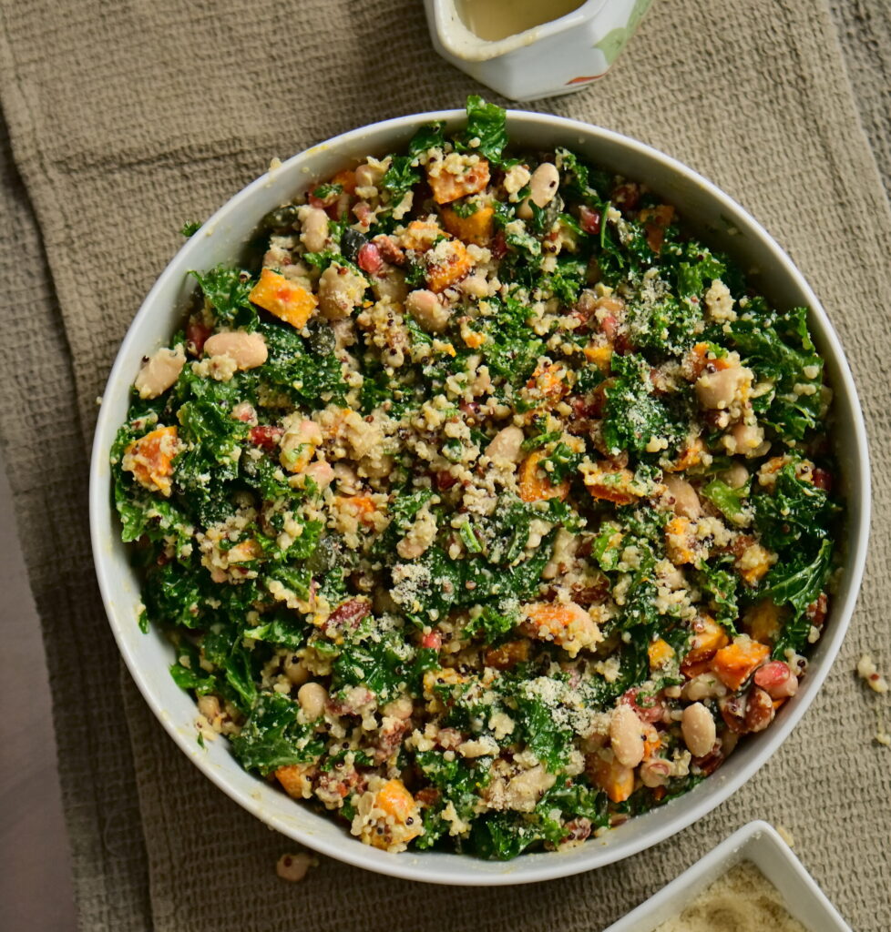 A serving bowl with kale and pumpkin salad, serving spoons, a pitcher, and almond flour on a brown tablecloth
