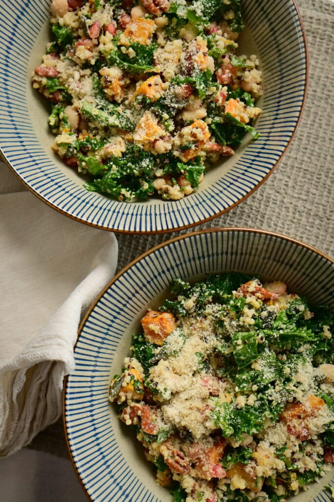 Two bowls with blue stripes filled with kale and pumpkin salad, on a brown tablecloth and a cream-colored cloth napkin.