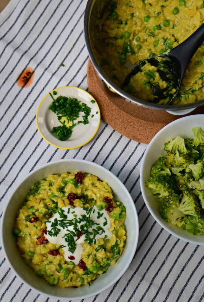 A messy table with a bowl of Savory ״Cheesy" Oatmeal, a bowl of chives, a bowl of broccoli, and a pot on a white cloth with blue stripes.