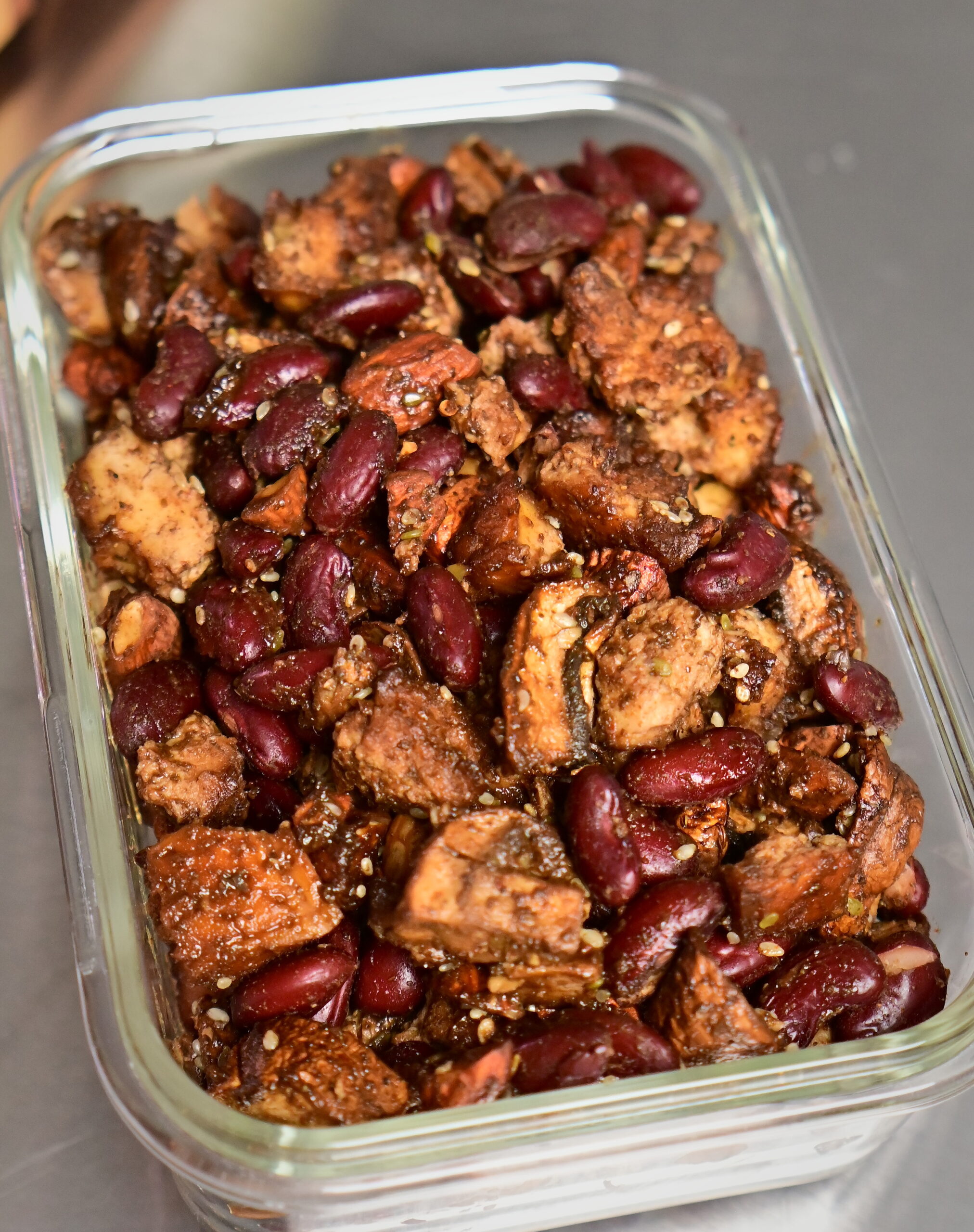 Protein mix of tofu, mushrooms, red beans, and seasoned baked almonds in a glass container on a metal surface.