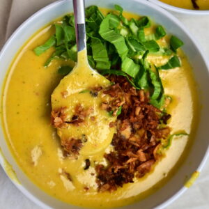 white bowl of creamy yellow potato soup with vegan bacon and greens on a white cloth, a spoon holding some soup.