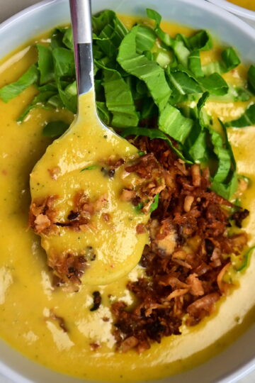 white bowl of creamy yellow potato soup with vegan bacon and greens on a white cloth, a spoon holding some soup.