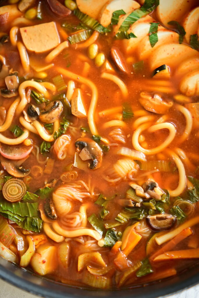close up to a vegan Korean Army Stew (Budae Jjigae) in a shallow pot on a white tablecloth.