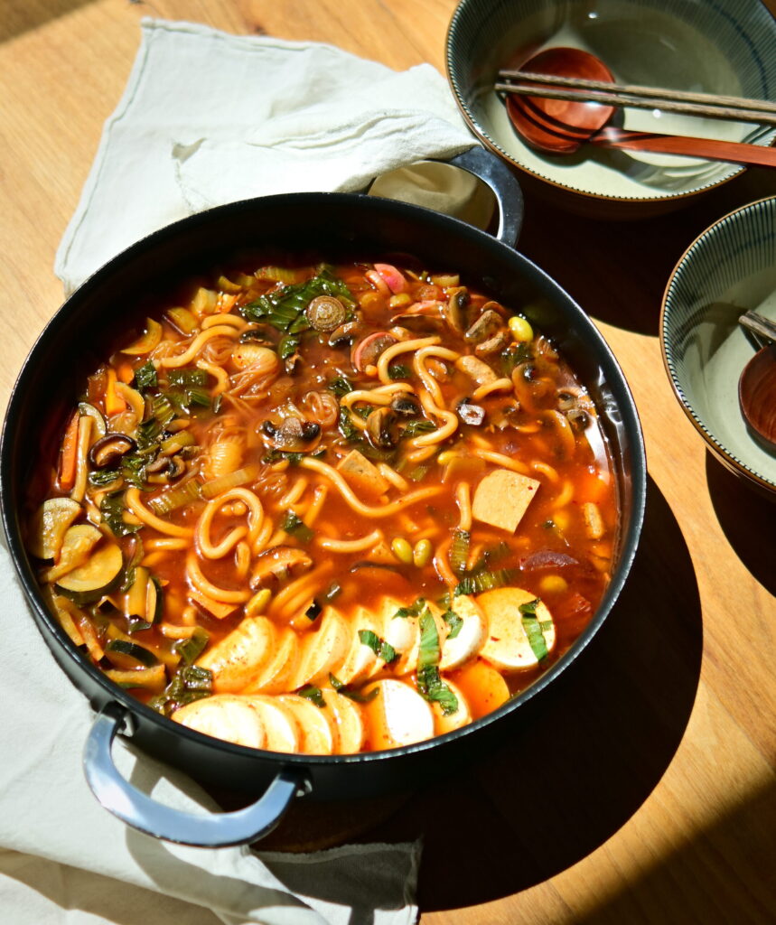 close up to a vegan Korean Army Stew (Budae Jjigae) in a shallow pot on a white tablecloth, with owls, chopsticks and spoons