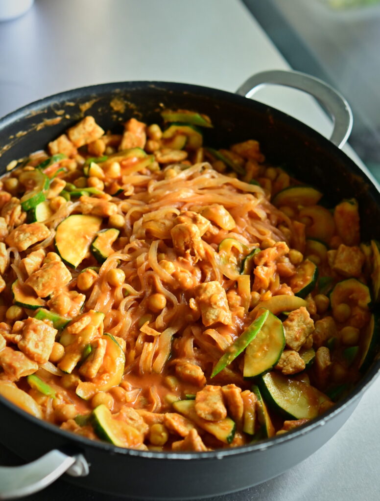 A wide, deep skillet filled with rosé noodles, chickpeas, zucchini, and vegan protein chunks on a metal countertop.