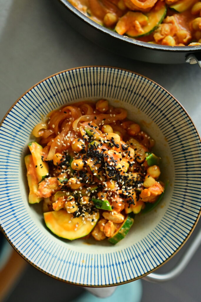 A striped deep bowl filled with rosé noodles, chickpeas, zucchini, and vegan protein chunks.